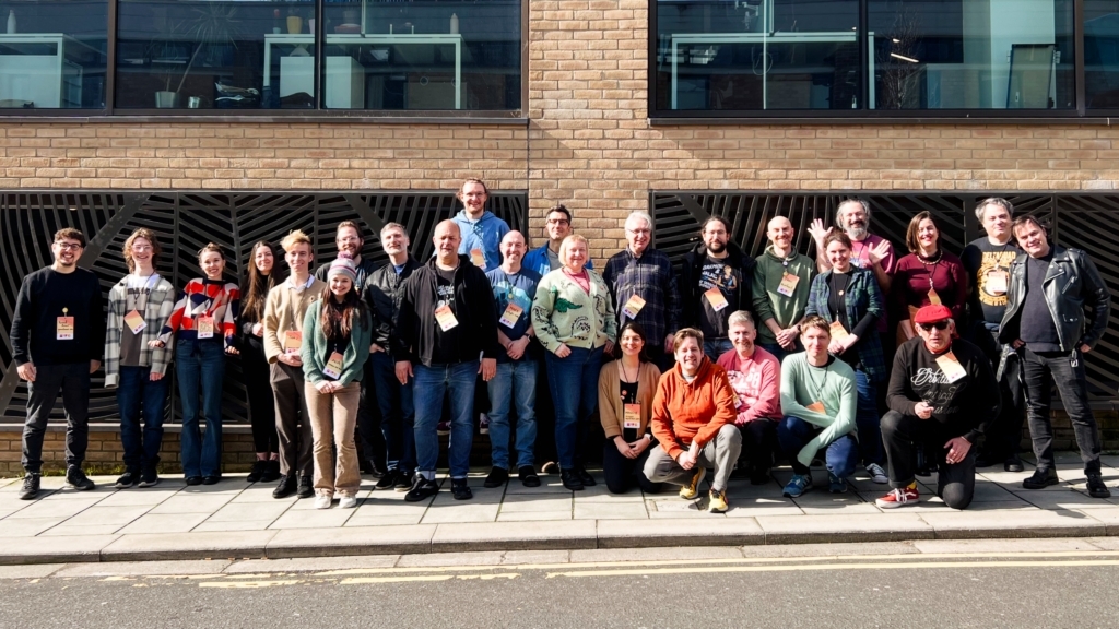 A group photo of the people that attended IndieWebCamp, including the author, taken outside the building against a brick wall in the sun. Everyone is smiling.