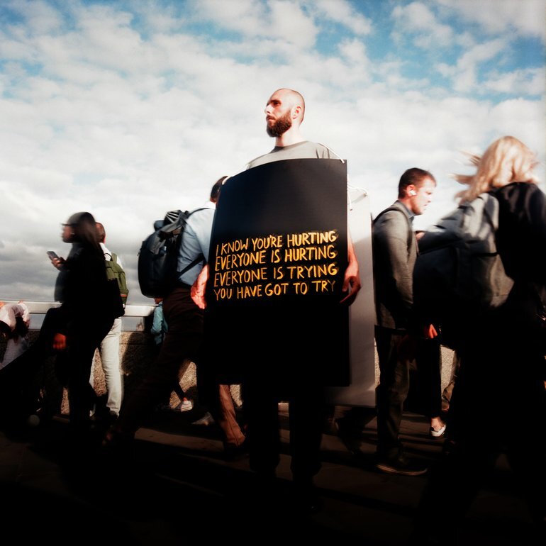 Cover art: A man stands stoically on a bridge as people rush past in all directions, wearing a large black placard with yellow handwritten text that reads: I know you're hurting, everyone is hurting, everyone is trying, you have got to try.