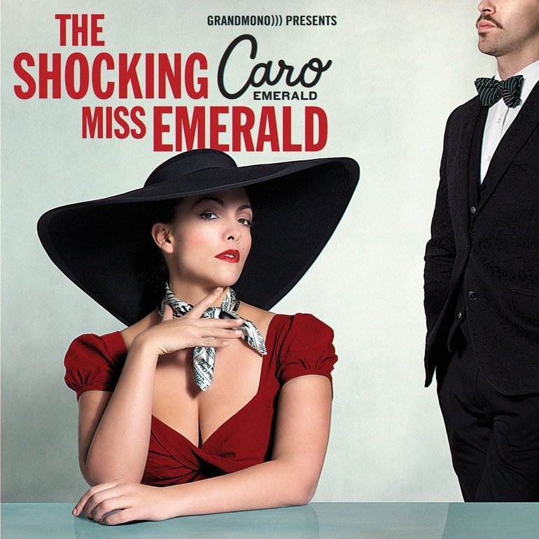 Cover art: Caro Emerald, wearing a wide-brimmed black hat, elegant newspaper print neckerchief, and revealing red dress, is sat at a glossy table, one arm lazily rested on its surface, the other hovering near her throat. She wears an expression of contempt, in the manner of film noir femme fatales. To her right is a man in a suit and bowtie, standing still and rigid facing towards her, but largely cut out of frame. Above is red text that reads: the shocking Miss Emerald, Caro Emerald.